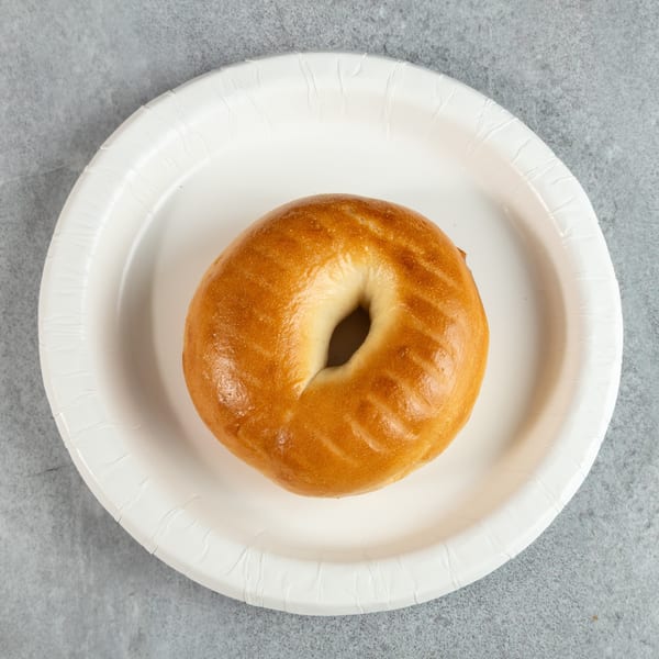 A plain, golden-brown bagel sits centered on a white paper plate against a gray background.