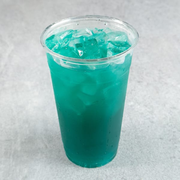 A clear plastic cup filled with a vibrant blue-green drink and ice cubes, on a gray surface.