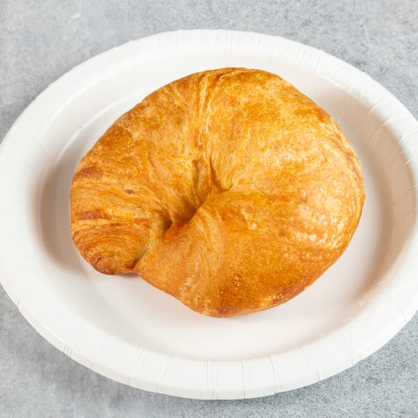 Golden-brown croissant on a white paper plate, centered on a gray textured surface.