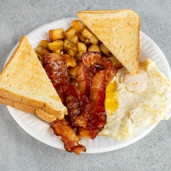 A breakfast platter with crispy bacon, fried eggs, home fries, and buttered toast on a white plate.