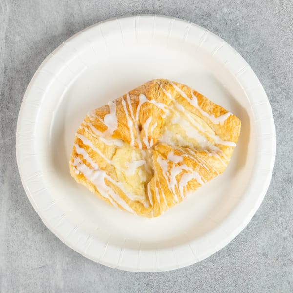 A square cheese danish with white icing drizzle sits on a white paper plate.